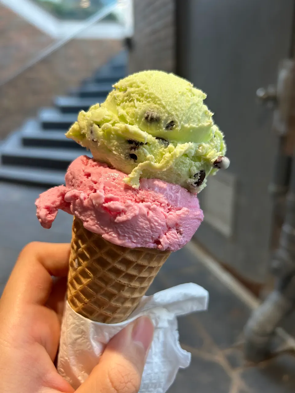 A hand holding a waffle cone with two scoops of ice cream: a pink scoop on the bottom and a light green scoop with chocolate chips on top, wrapped in a white napkin.