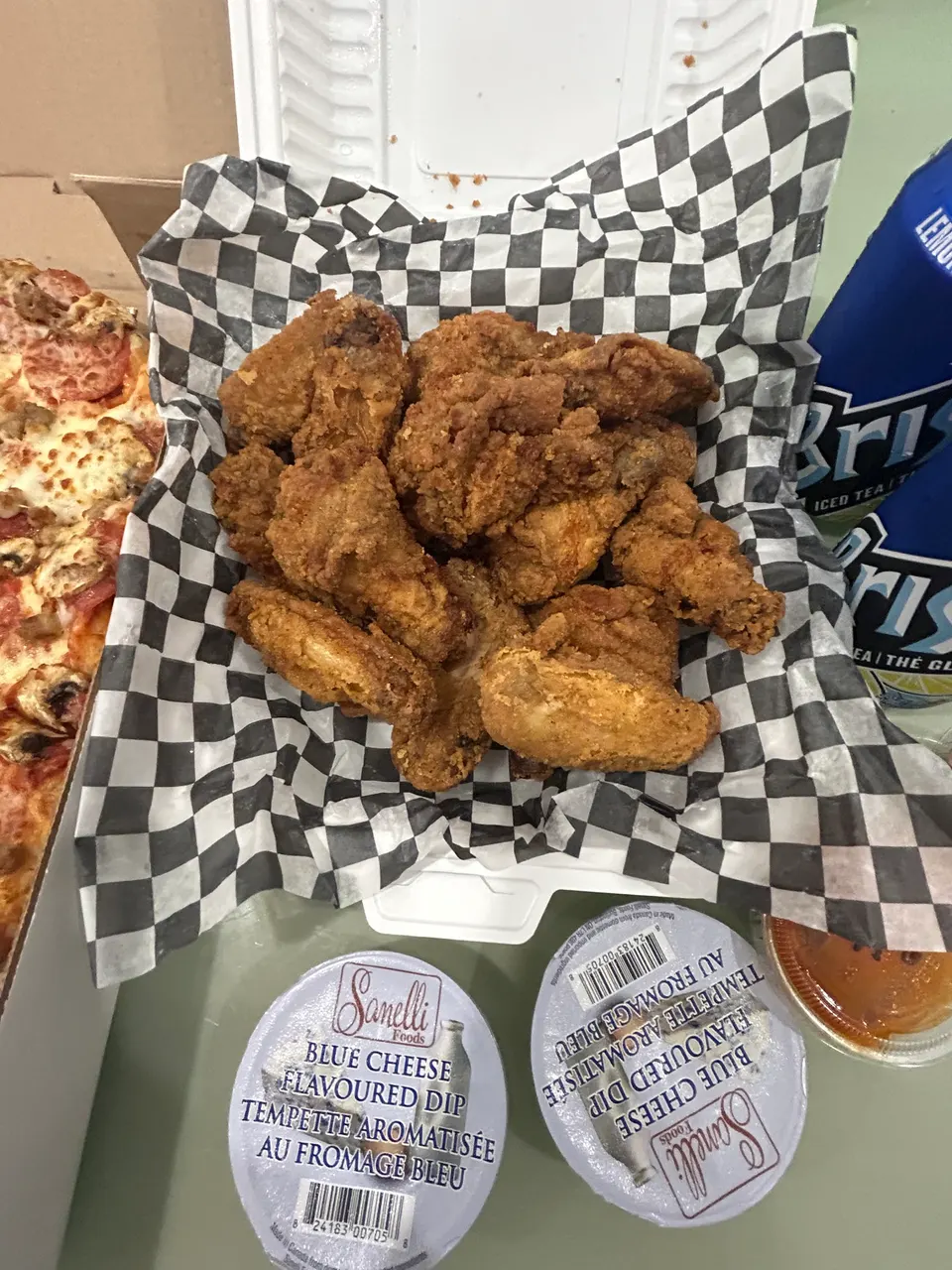 A basket of crispy golden-brown chicken wings served on black and white checkered paper, next to a pepperoni pizza, two containers of Sanelli blue cheese dip, and a blue can of Brisk iced tea.