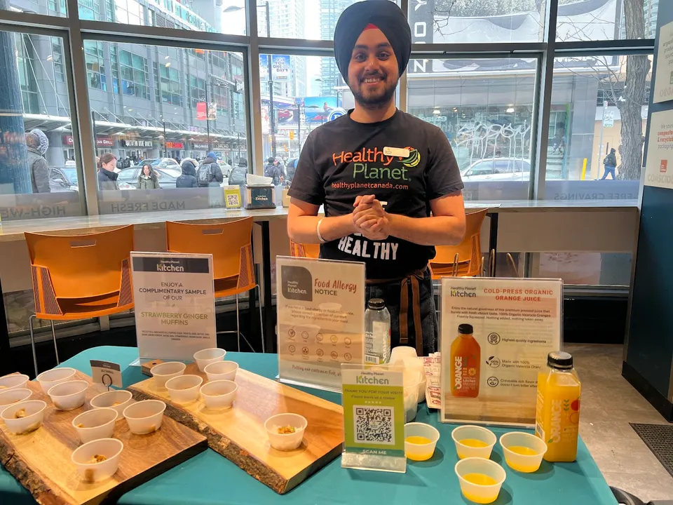 A smiling staff member wearing a turban and a Healthy Planet t-shirt stands behind a sampling table with small cups of orange juice and muffin samples on wooden boards.