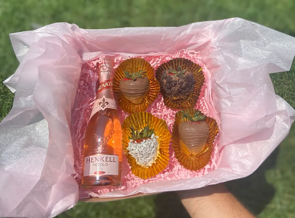 A gift box lined with pink tissue paper containing a small bottle of Henkell Piccolo rosé sparkling wine and four gourmet chocolate-covered strawberries in gold liners.