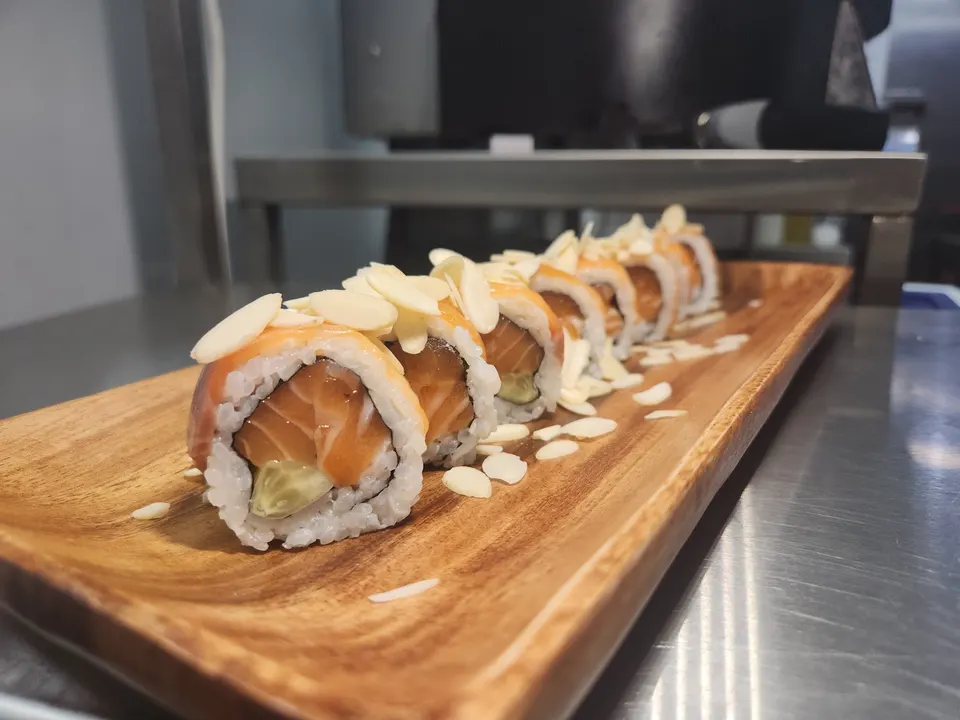 A row of salmon sushi rolls topped with sliced almonds, served on a long wooden platter on a stainless steel counter.