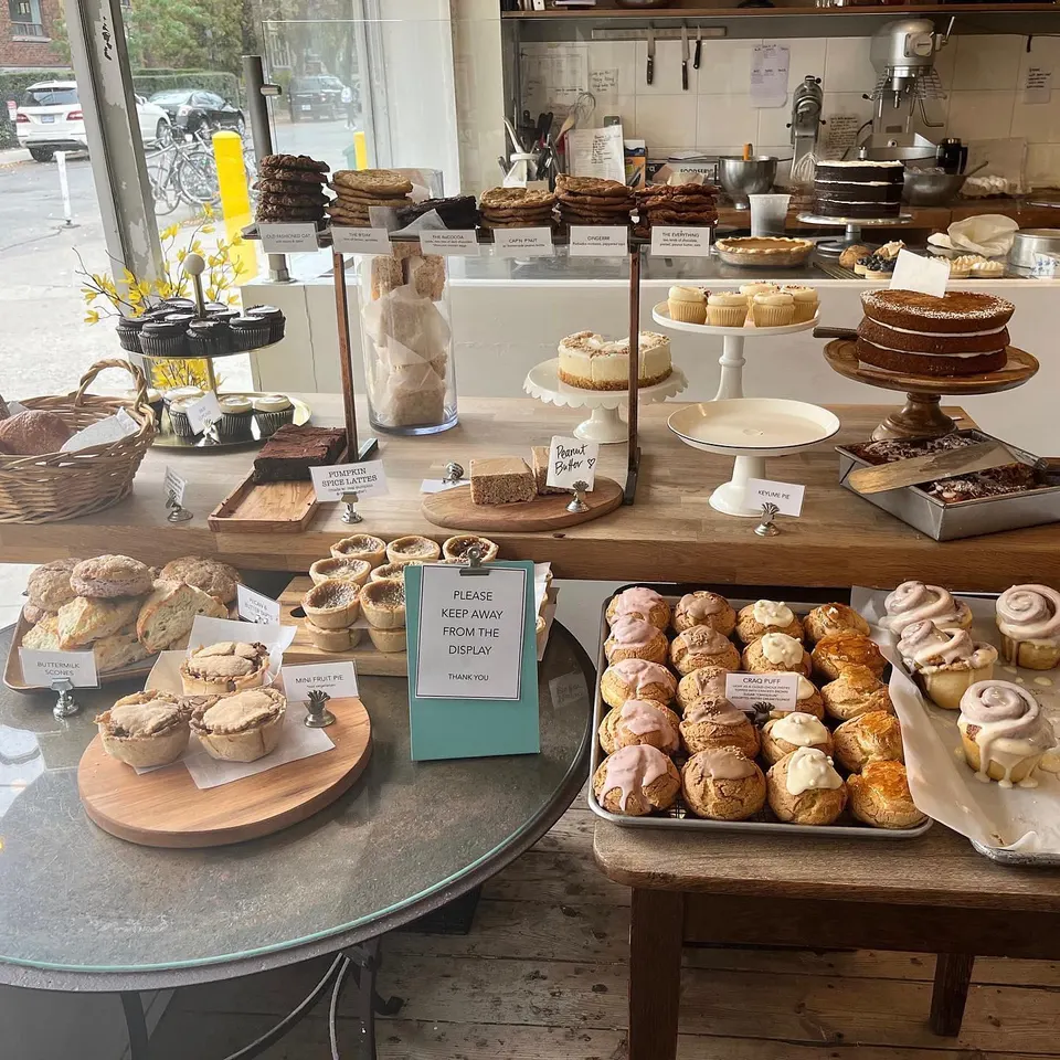 A bakery display counter filled with various baked goods including cookies, cakes, cupcakes, tarts, scones, cream puffs, and cinnamon rolls on wooden stands and trays.