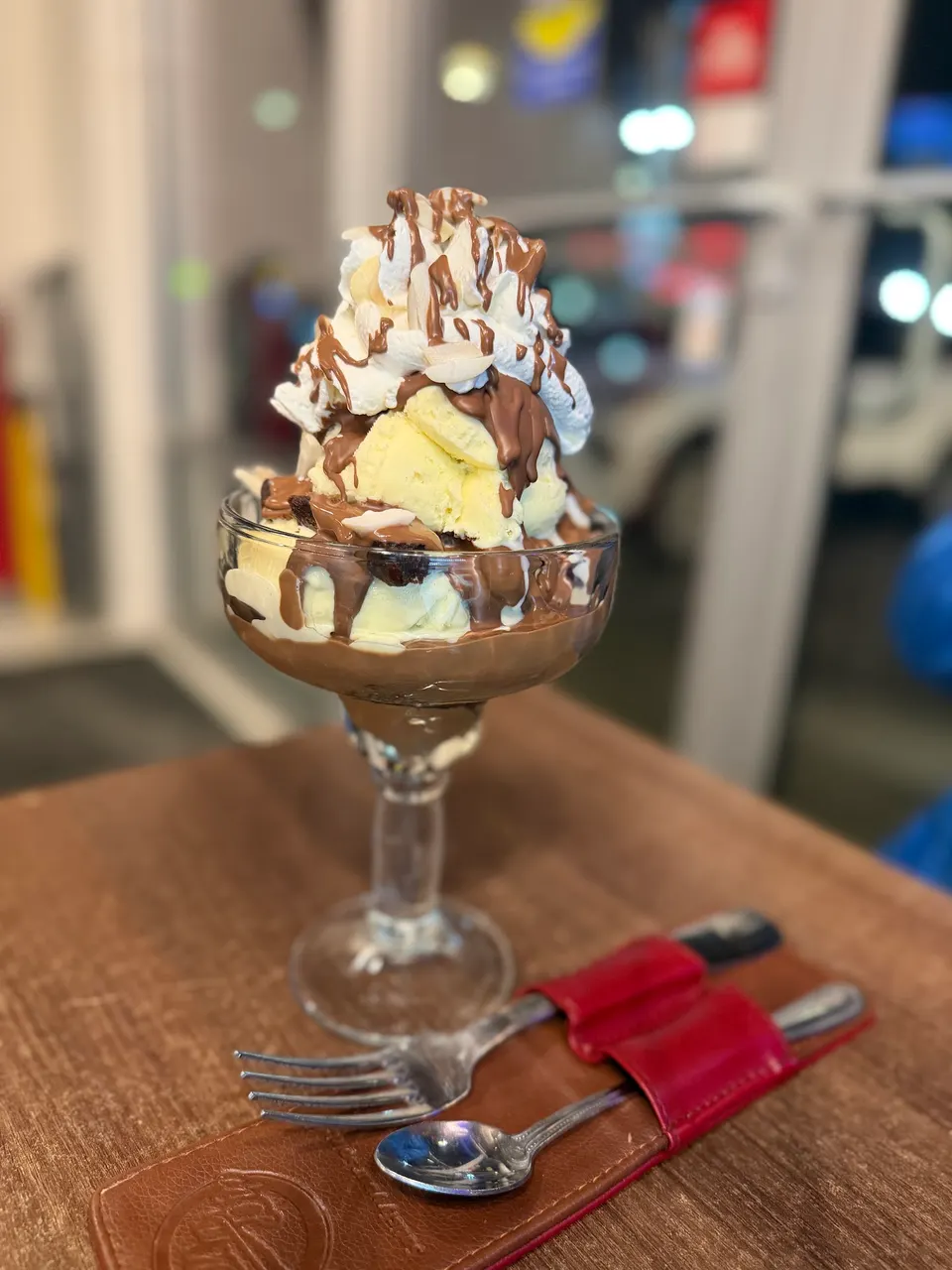 A large ice cream sundae in a glass goblet, topped with vanilla ice cream, whipped cream, chocolate sauce, and sliced almonds, served on a wooden table with a fork and spoon.