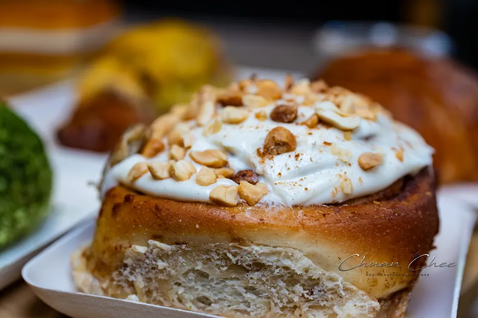 A close-up of a freshly baked cinnamon roll topped with creamy white frosting and sprinkled with chopped peanuts, served on a small white tray.