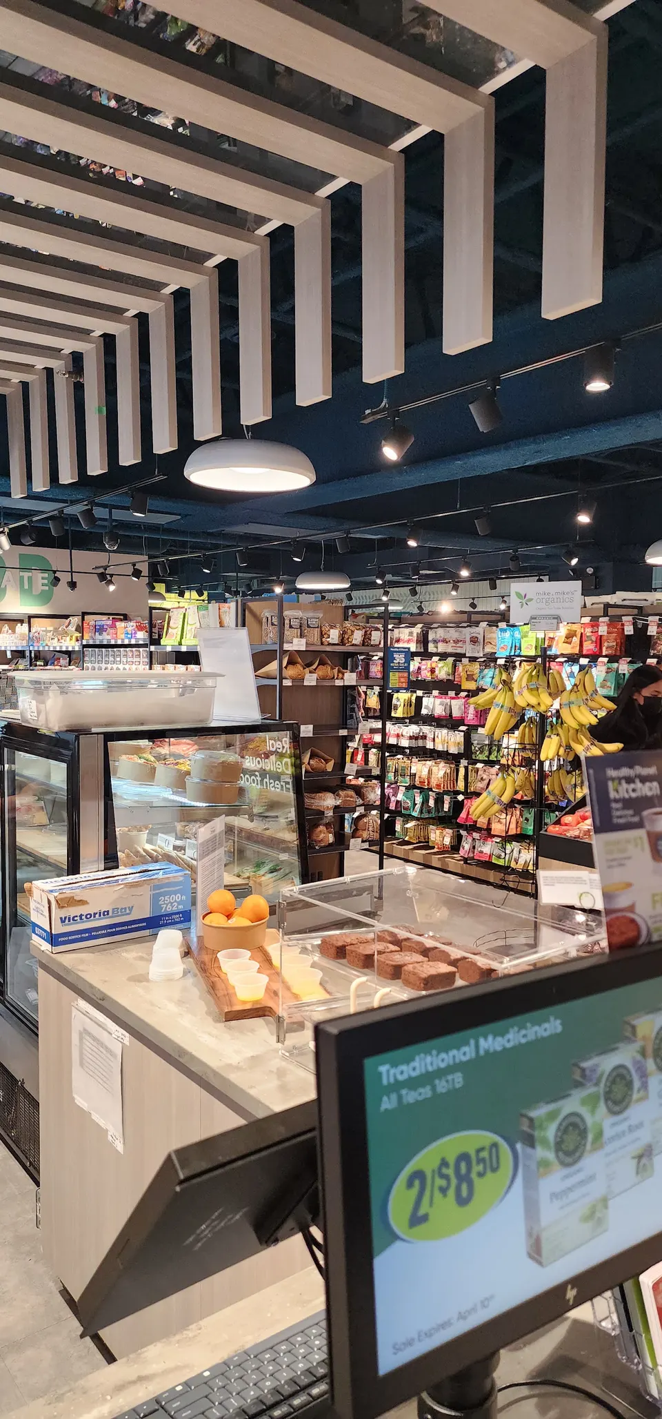 A checkout counter at Healthy Planet Kitchen featuring a display of baked goods, fresh oranges, and a digital screen showing a promotion for Traditional Medicinals teas.