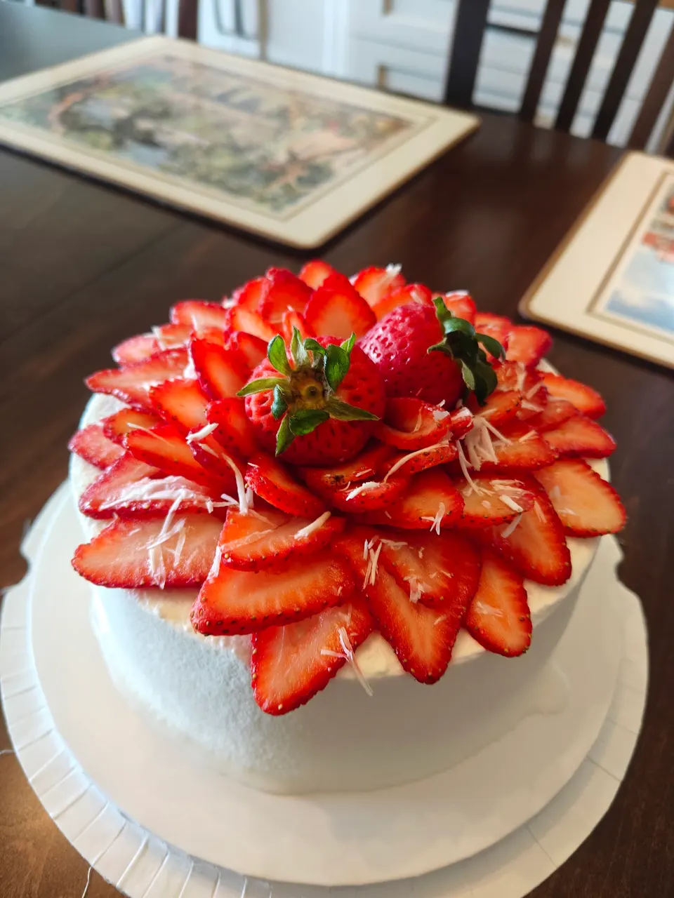 A round white frosted cake topped with concentric layers of sliced fresh strawberries and two whole strawberries with green stems in the center.