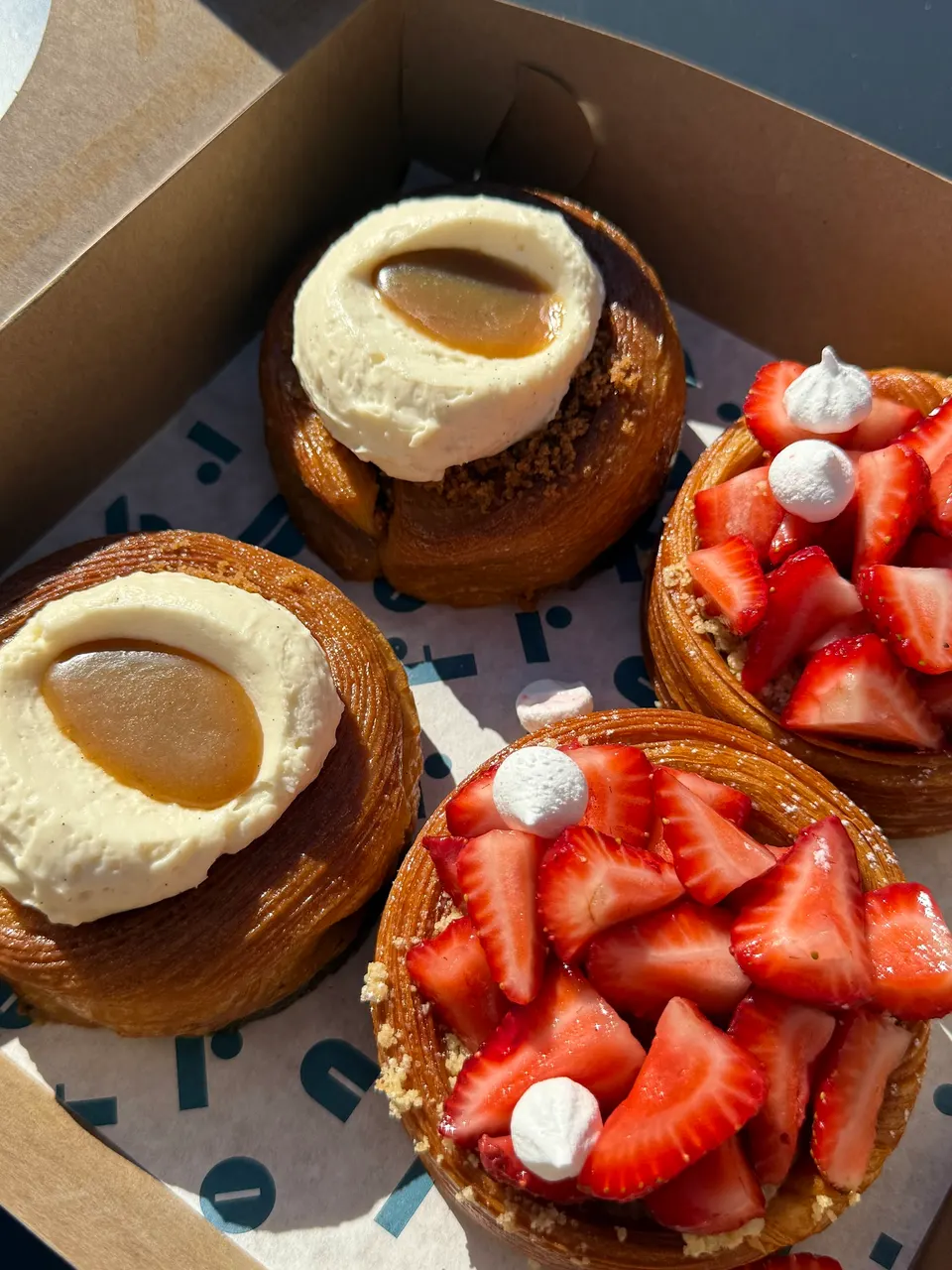 Four round pastries in a cardboard box, two topped with white cream and caramel, and two topped with sliced fresh strawberries and small white meringues.