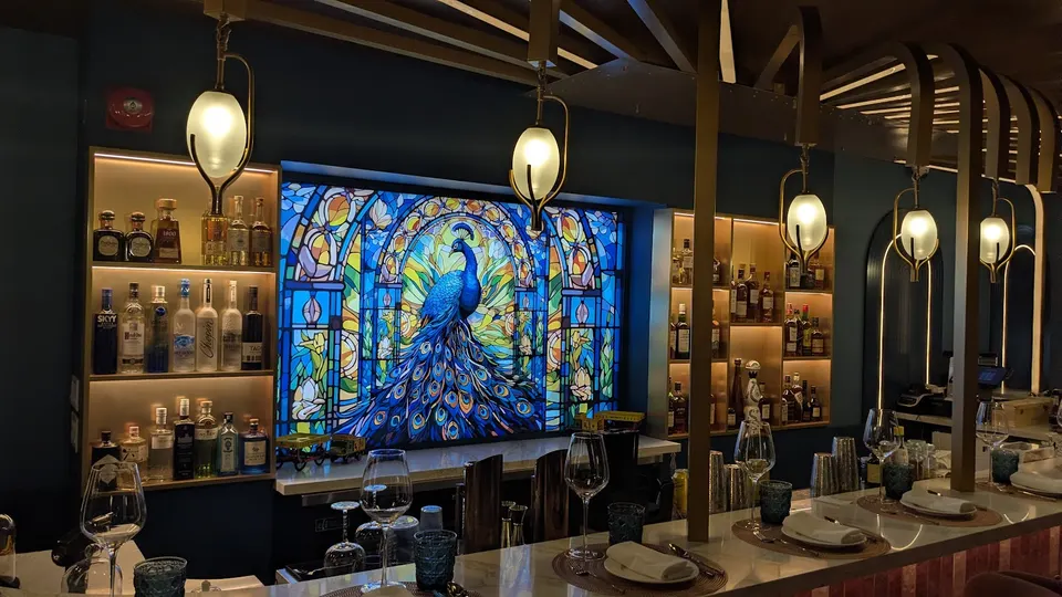 A stylish bar counter at Aangan Toronto featuring a vibrant stained-glass window depicting a peacock, flanked by illuminated liquor shelves and elegant hanging pendant lights.