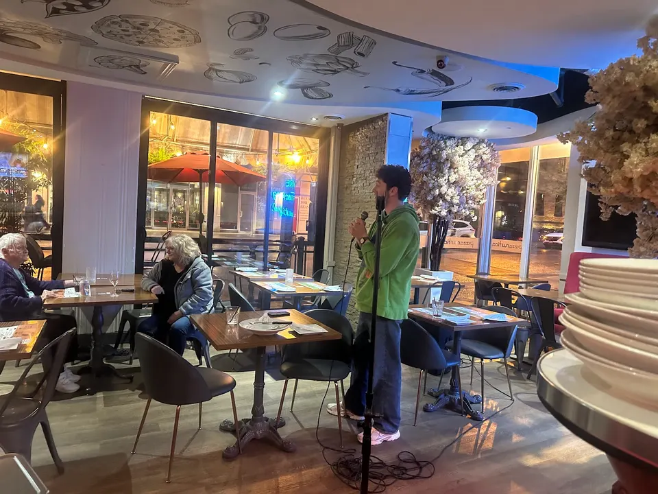 A man in a green hoodie stands holding a microphone, performing in the dining area of Pizza Rustica. Customers sit at nearby wooden tables, and a stack of plates is visible in the foreground.