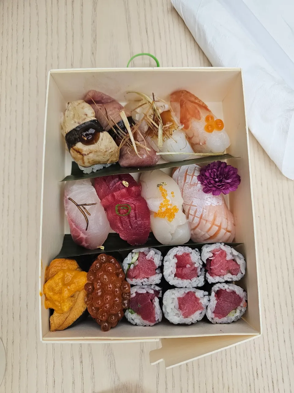 A wooden bento box filled with an assortment of premium sushi, including nigiri topped with uni, ikura, tuna, and shrimp, alongside several tuna maki rolls.
