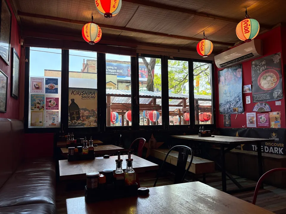 Interior dining area of Tondou Ramen featuring wooden tables, leather booths, and metal chairs under hanging Japanese paper lanterns, with large windows looking out.