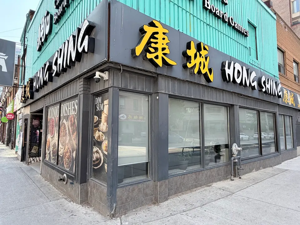 Exterior view of Hong Shing Restaurant on a city street corner, featuring large black and yellow signage with Chinese characters and English text, large windows, and food posters on the side wall.