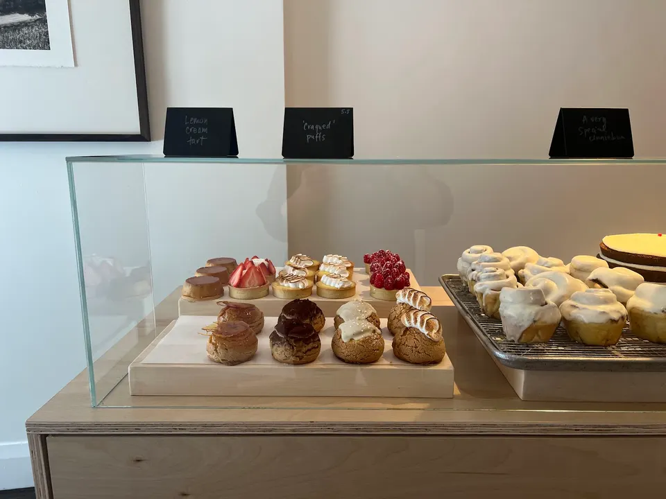 A glass display case at Bakerbots Baking filled with various pastries, including cream puffs, fruit tarts, and iced cinnamon rolls on wooden and metal trays.