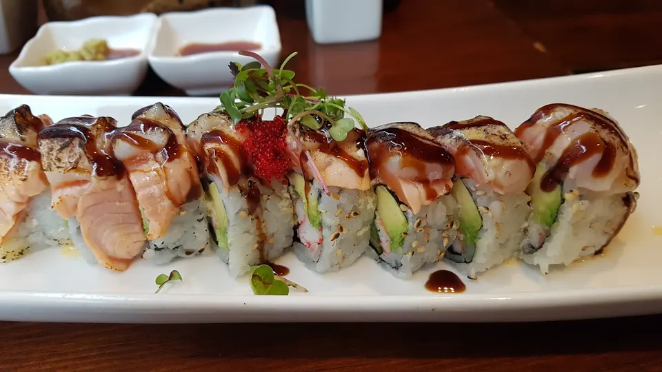 A close-up shot of a specialty sushi roll topped with seared salmon, dark sweet sauce, microgreens, and red tobiko, served on a long white plate with small dipping bowls in the background.