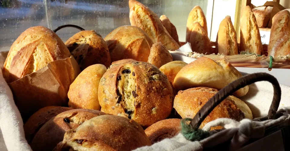 A close-up shot of a rustic basket filled with an assortment of freshly baked artisanal breads, including round loaves, baguettes, and seeded buns, bathed in warm sunlight.