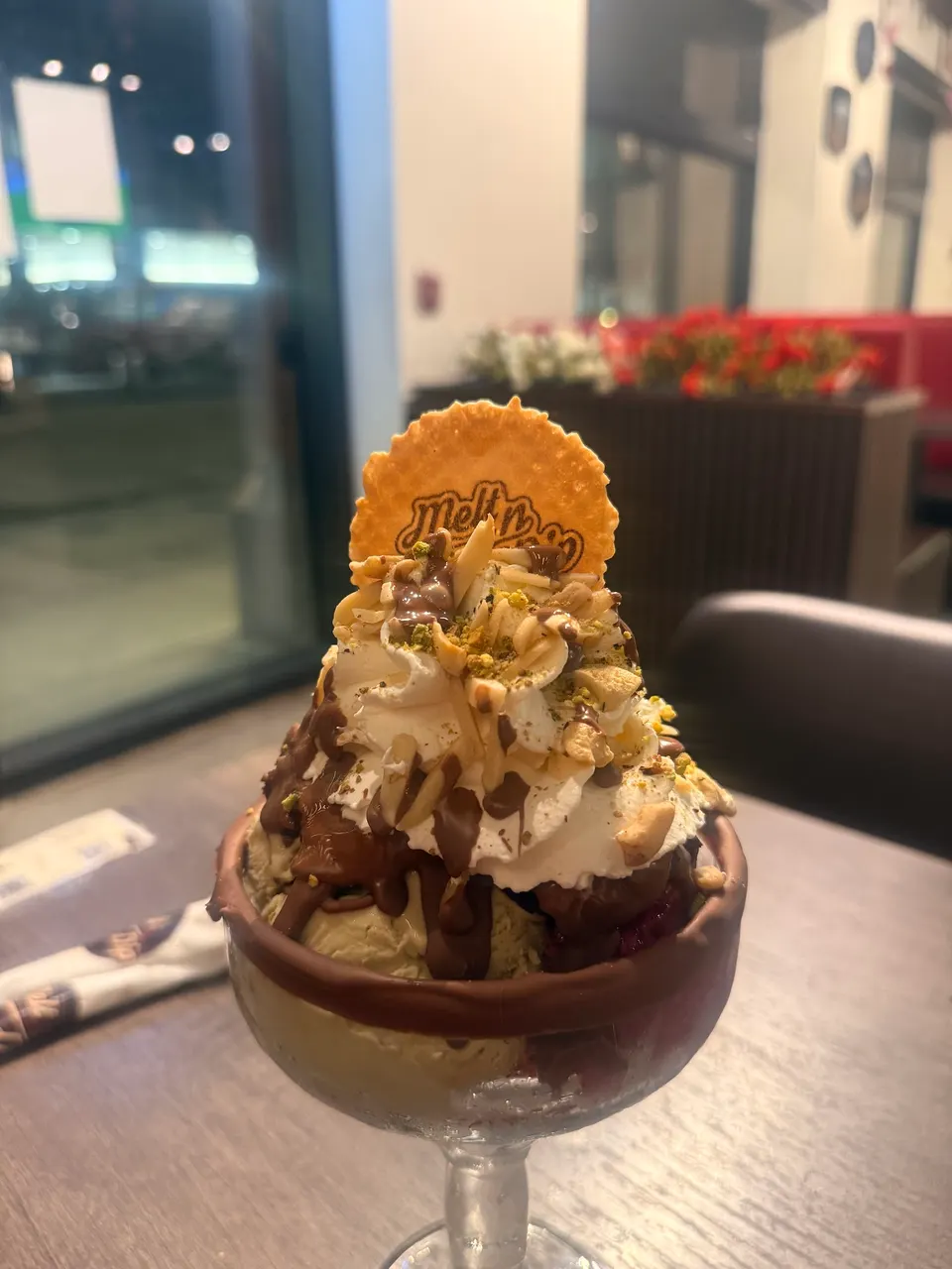 A decadent ice cream sundae served in a glass goblet, topped with whipped cream, chocolate sauce, chopped nuts, and a round waffle cookie with the 'Melt n dip' logo.
