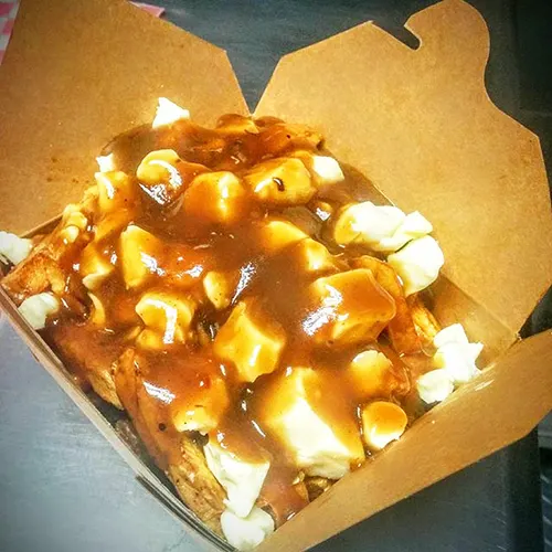A close-up shot of a poutine dish served in a brown cardboard takeout container, featuring thick-cut french fries topped with white cheese curds and rich brown gravy.