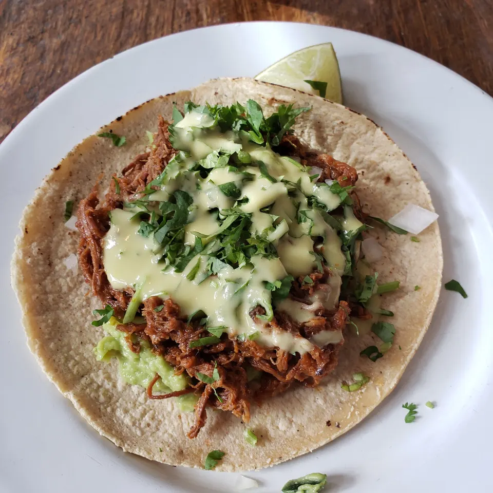 A single taco served on a soft corn tortilla, topped with shredded beef, guacamole, chopped cilantro, onions, and a creamy green salsa, with a lime wedge on the side.