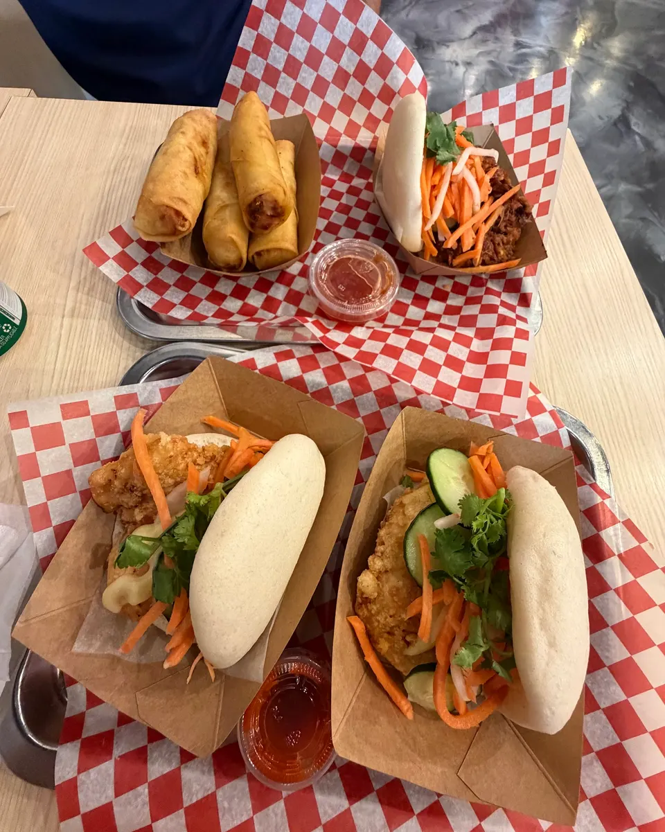 Three cardboard trays on metal platters with red and white checkered paper liners, containing fried spring rolls, a pulled pork bao, a fried chicken bao, and a crispy fish bao with pickled vegetables.