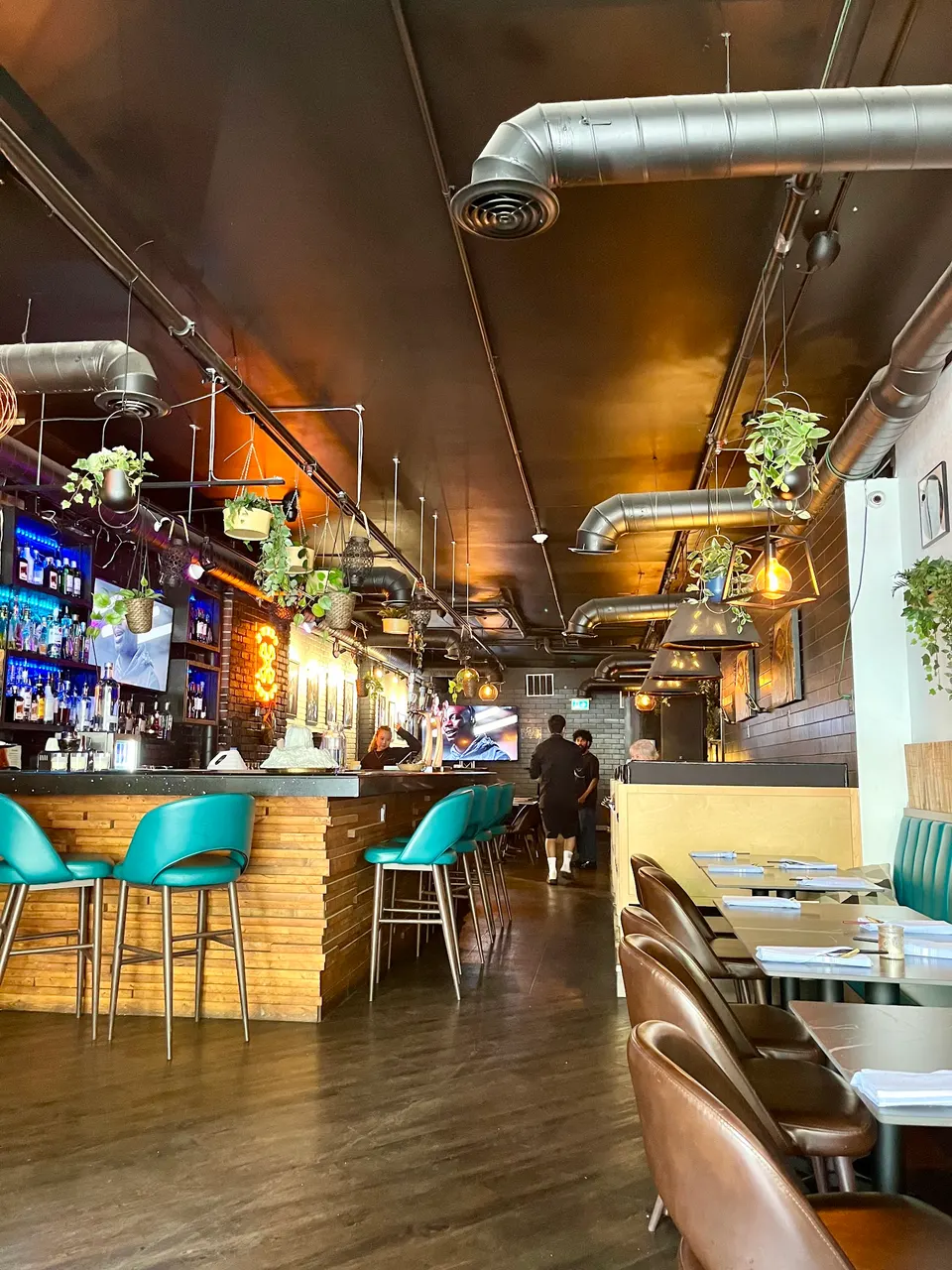 Interior view of Crystal Thani Kitchen & Bar showing a wooden bar counter with teal stools on the left, dining tables with brown leather chairs on the right, and hanging plants under a dark ceiling with exposed ductwork.