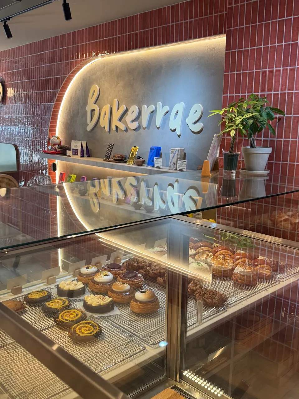 A glass display case filled with various pastries and baked goods inside Bakerrae bakery, with a large 'Bakerrae' sign on a grey wall in the background.