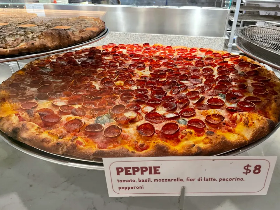 A large, freshly baked pepperoni pizza with curled, crispy pepperoni slices on a metal tray, displayed next to a menu card labeled 'Peppie' for $8.