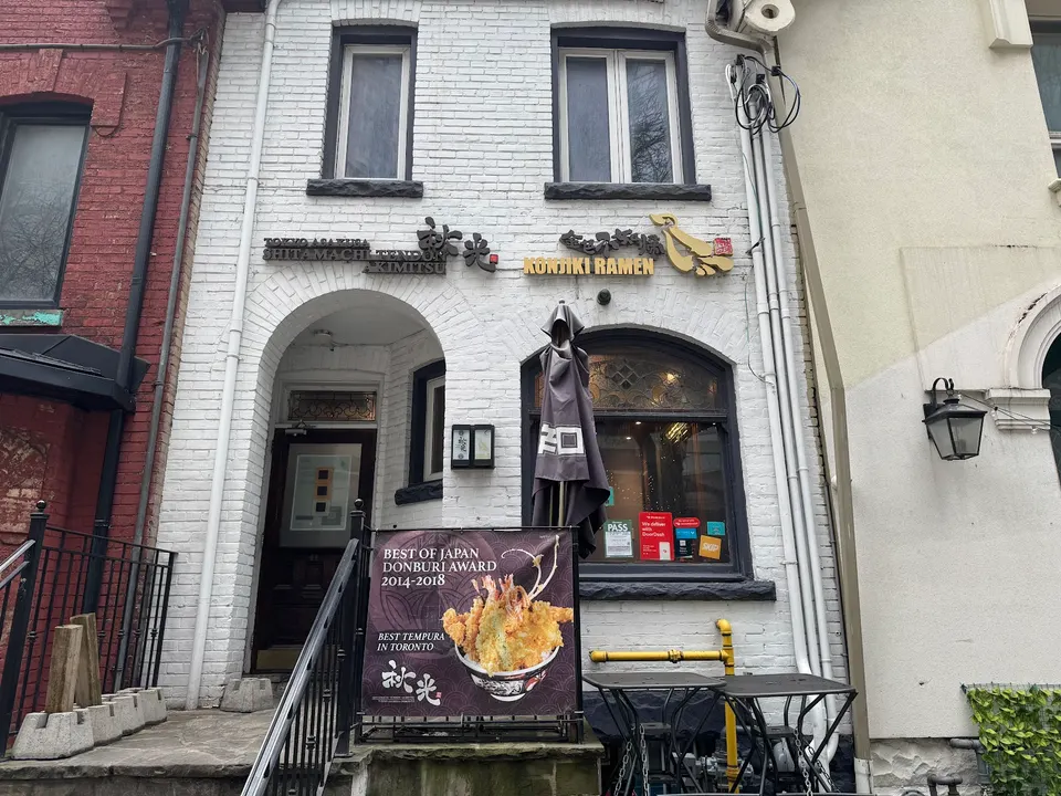 Exterior of Konjiki Ramen in Downtown Toronto, featuring a white brick facade, two signs for the restaurant, a black arched window, and a promotional sign for tempura awards.
