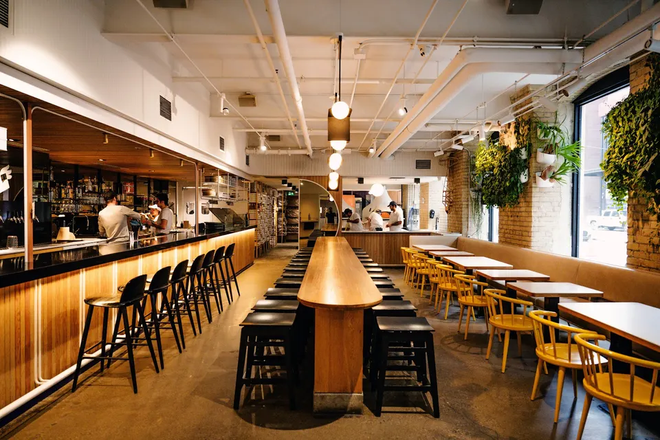 Interior of General Assembly Pizza showing a long wooden communal table, bar seating with black stools, yellow dining chairs, and hanging plants on brick walls.