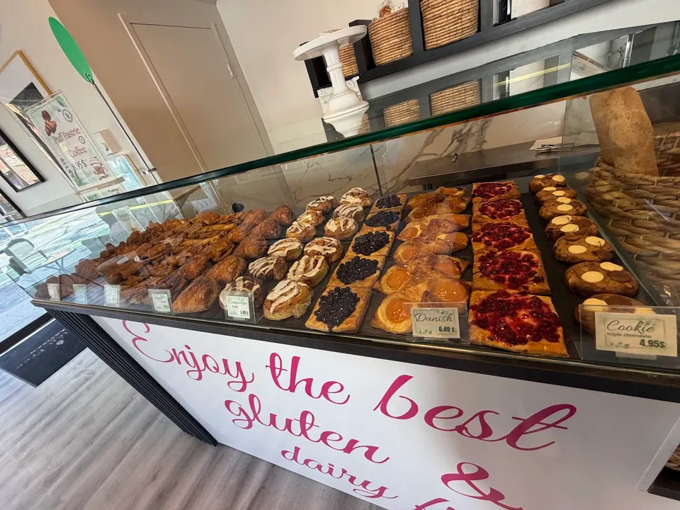 A bakery display case filled with rows of gluten-free pastries, including danishes with fruit toppings, cinnamon rolls, and chocolate chip cookies, with a sign reading 'Enjoy the best gluten & dairy free'.