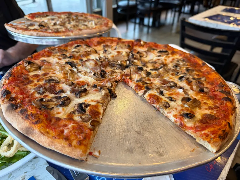 A large, thin-crust pizza with mushrooms and melted cheese, with a few slices missing, served on a metal tray at Cafe Diplomatico Restaurant & Pizzeria. Another whole pizza is visible on a raised stand in the background.