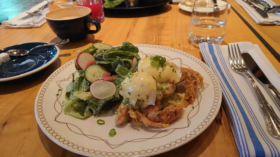 A plate of soft-shell crab eggs benedict topped with hollandaise sauce and green onions, served with a side salad of lettuce, cucumber, and radishes on a wooden table.