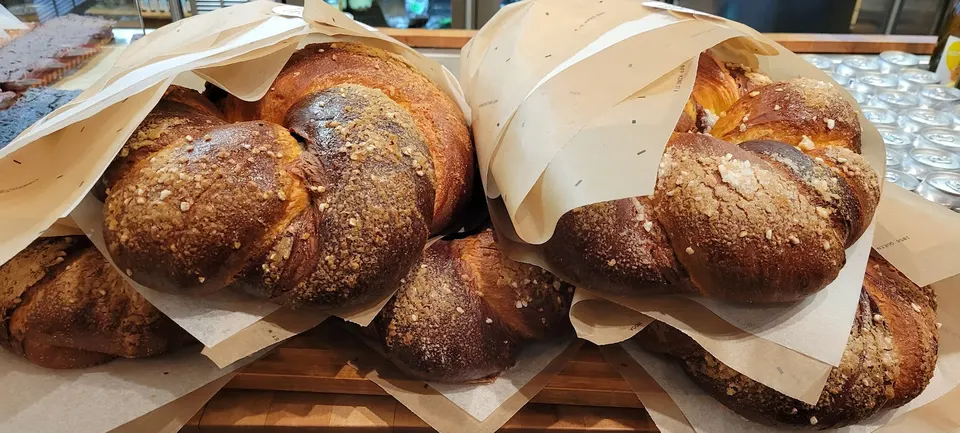 Large, braided brioche loaves with a golden-brown crust and sugar topping, wrapped in parchment paper on a wooden bakery display shelf.