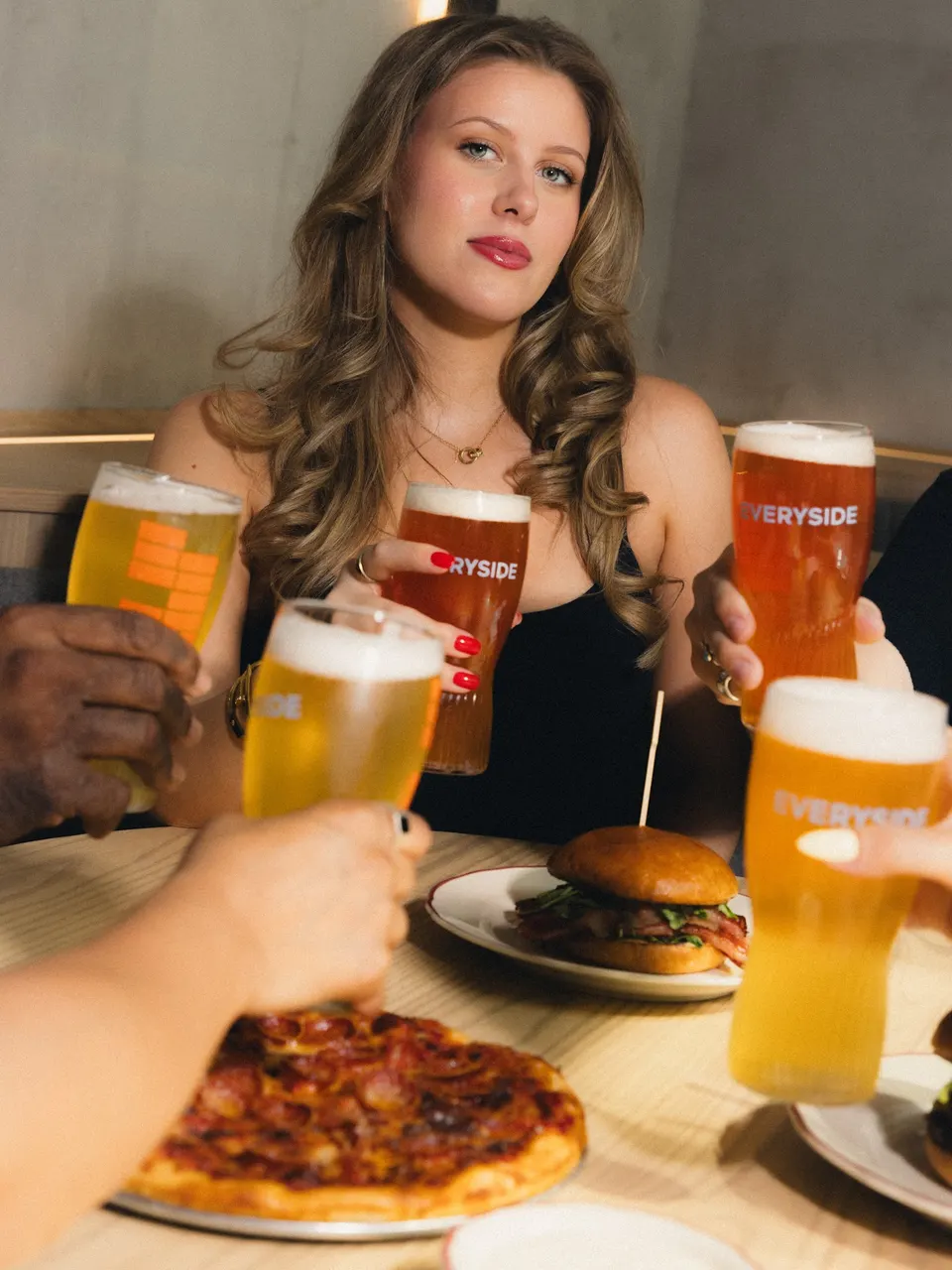 A group of friends toast with glasses of beer over a table featuring a pepperoni pizza and a burger at Everyside - Social Eatery & Taphouse.