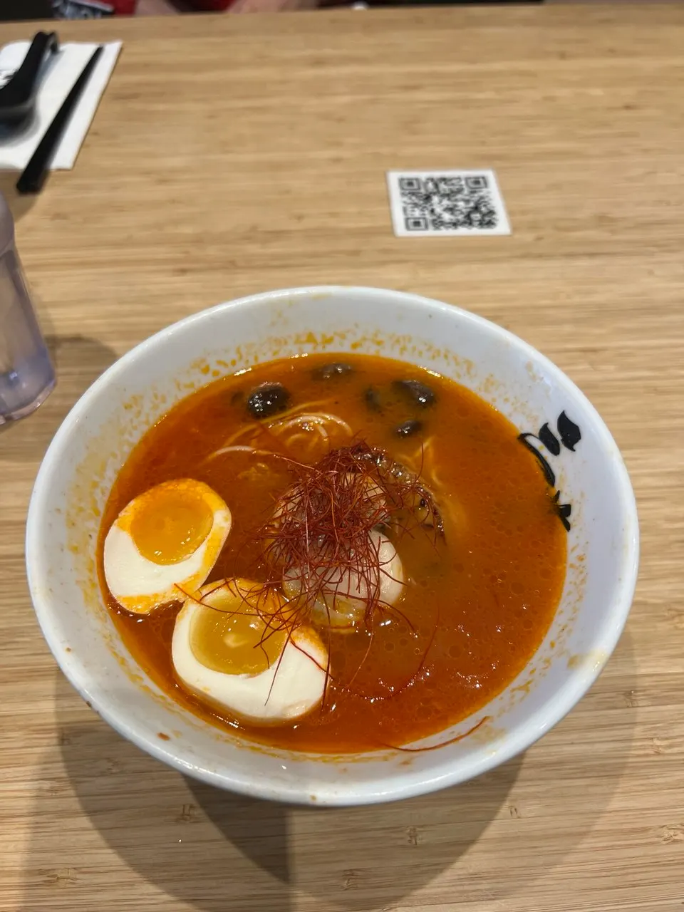 A bowl of spicy ramen with a rich red broth, soft-boiled egg halves, noodles, and red chili threads on top, served in a white bowl on a wooden table.