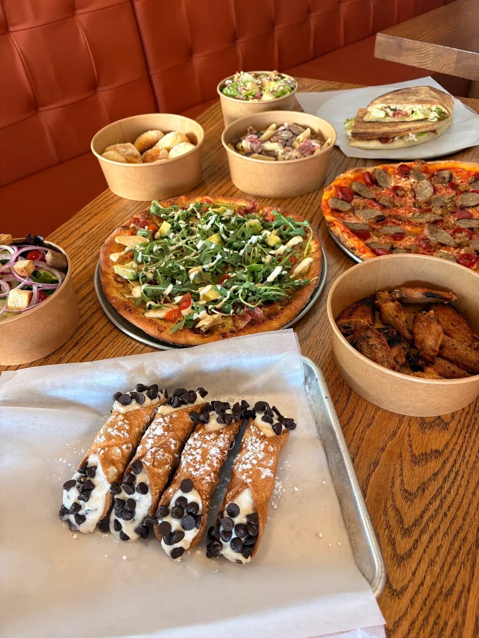 A wooden table spread with a variety of Italian dishes including two wood-fired pizzas, a bowl of pasta, chicken wings, garlic bites, salads, a panini, and four chocolate chip cannoli on a tray in the foreground.