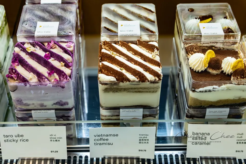 Three varieties of square mini cakes in clear plastic containers displayed in a bakery case, including Taro Ube Sticky Rice, Vietnamese Coffee Tiramisu, and Banana Salted Caramel.