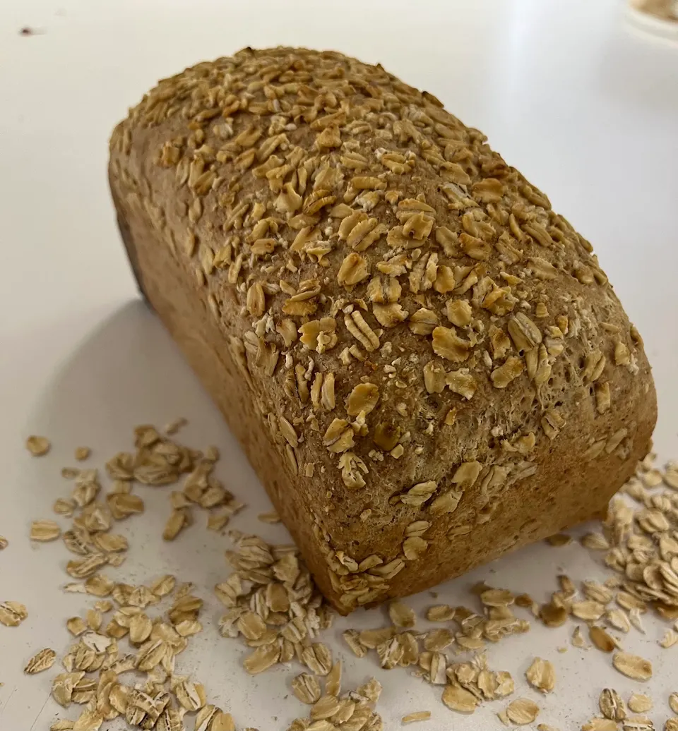 A freshly baked loaf of whole wheat bread topped with rolled oats, sitting on a white surface with loose oats scattered around its base.