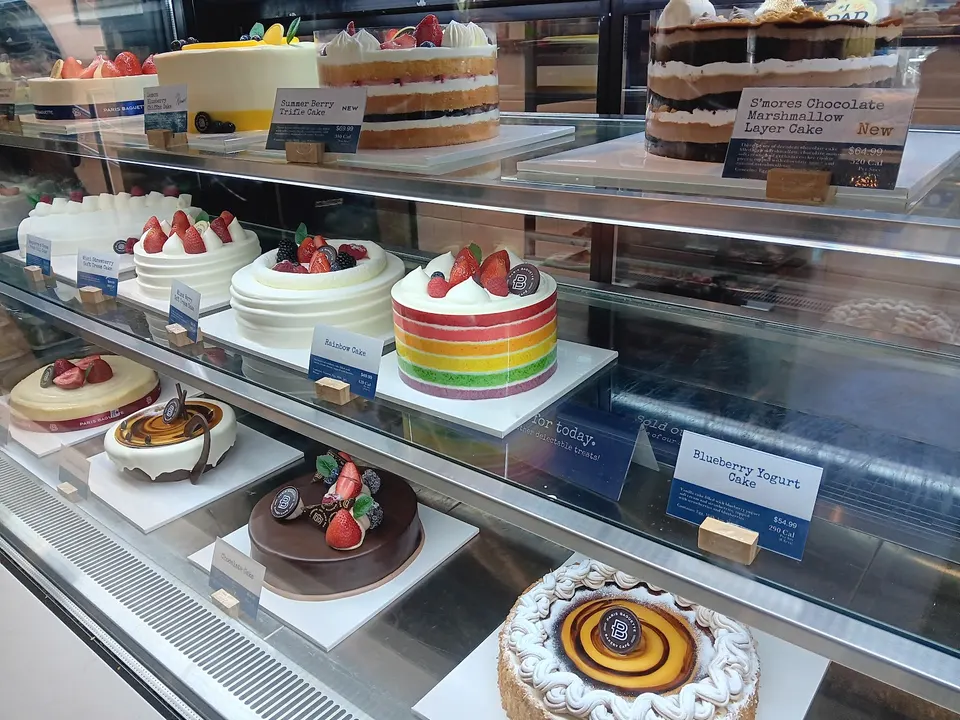 A glass display case at Paris Baguette filled with various decorated whole cakes, including a rainbow cake, chocolate cakes, and fruit-topped sponge cakes with price tags.