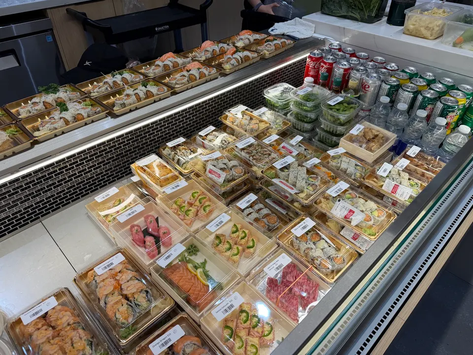 A refrigerated display case filled with pre-packaged sushi rolls, nigiri, sashimi, and side dishes in clear plastic containers, alongside canned sodas and bottled water.