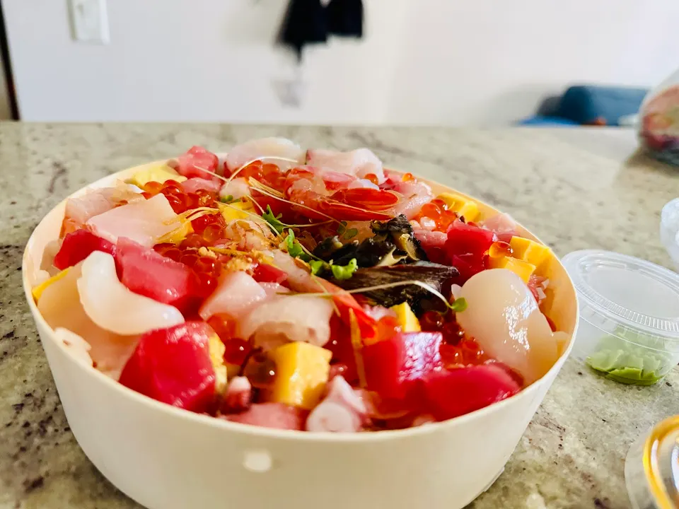A round wooden takeout box filled with chirashi sushi, featuring assorted diced raw fish, octopus, shrimp, salmon roe, and tamago on a granite countertop.