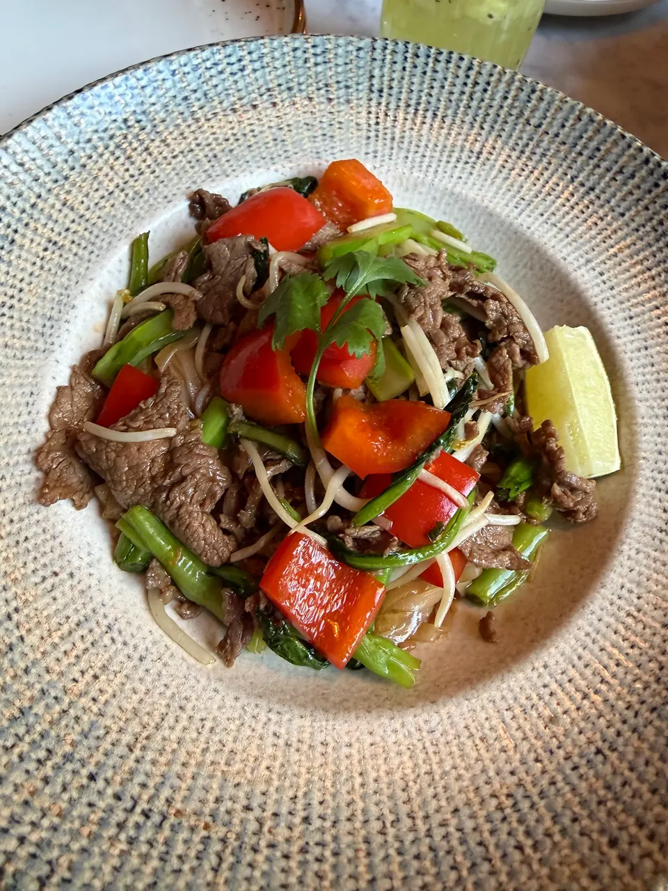 A stir-fried beef dish served in a textured ceramic bowl, featuring sliced beef, red bell peppers, green vegetables, and bean sprouts, garnished with cilantro and a lime wedge on the side.