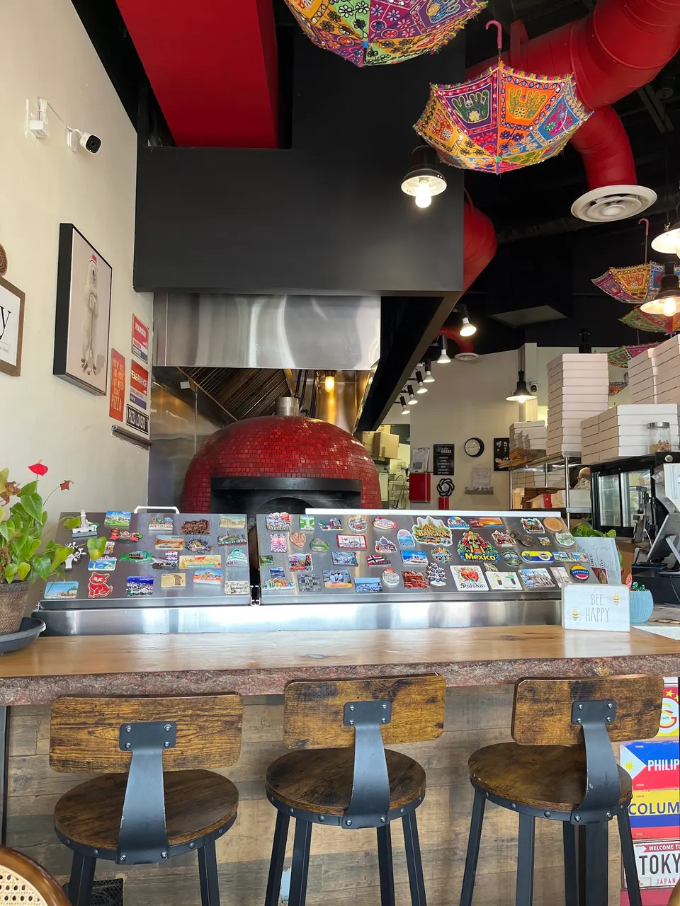 A wooden counter with three rustic bar stools in front of a red tiled pizza oven. Colorful umbrellas hang from the ceiling, and the counter is decorated with travel magnets.