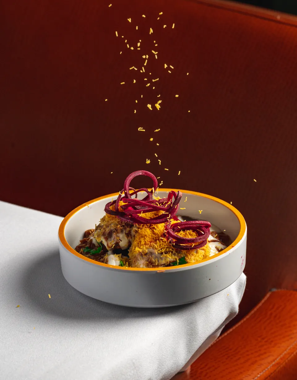 A bowl of Indian chaat topped with yellow sev and curled beetroot slices, with fine sev being sprinkled from above against a dark background.