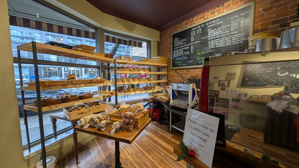 Interior of a bakery showing wooden shelves stocked with fresh bread and pastries, a small display table with packaged baked goods, a menu board on a brick wall, and a service counter.