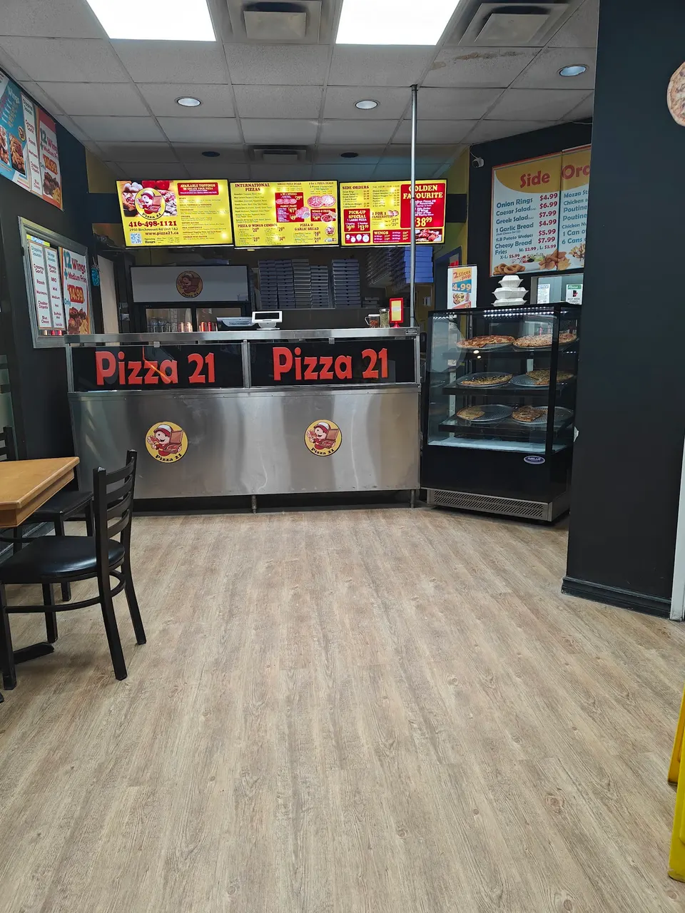 Interior of Pizza 21 showing the ordering counter with stainless steel panels, digital menu boards overhead, a glass display case with pizzas, and a dining table with chairs in the foreground.