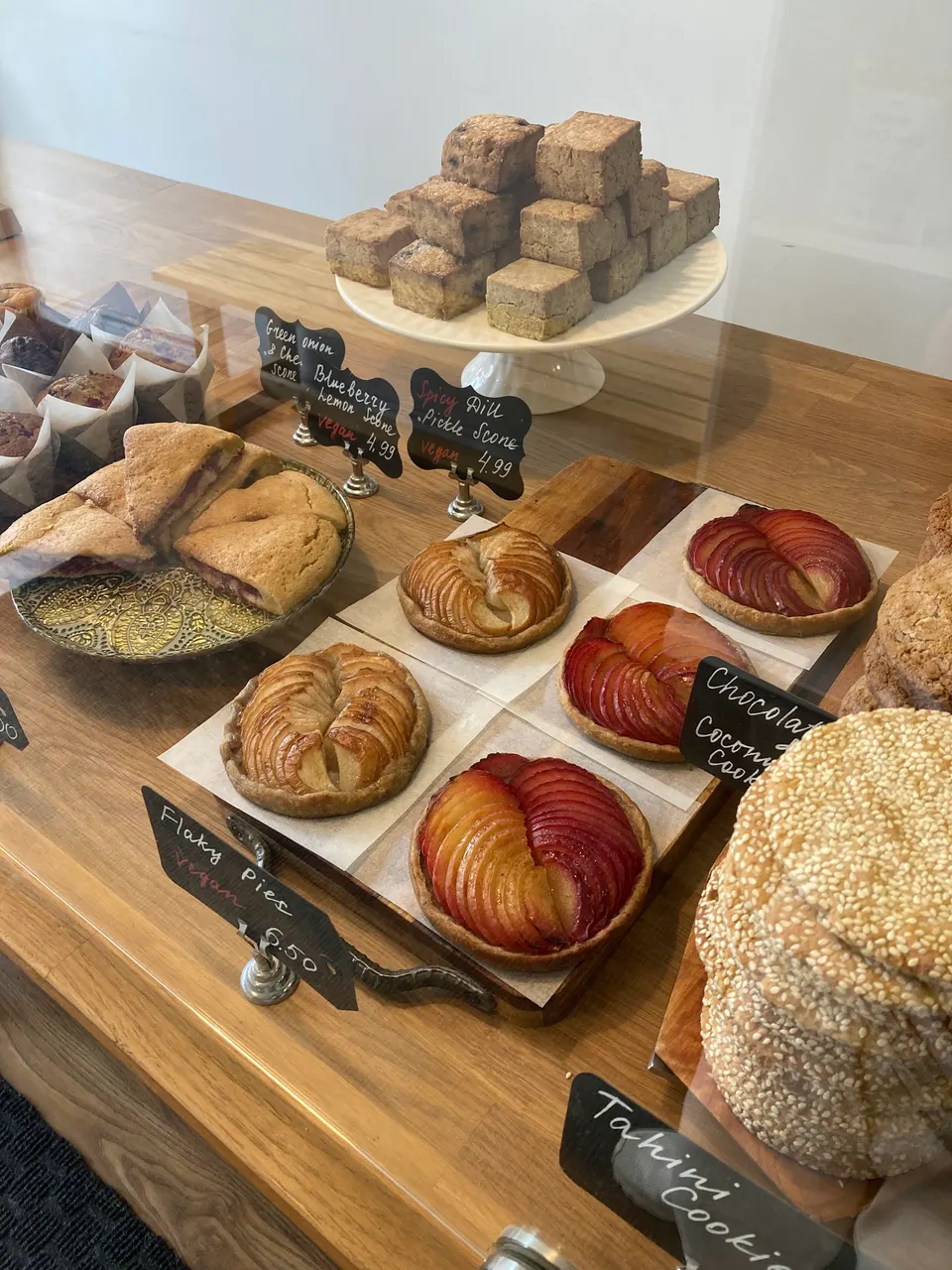 A bakery display counter featuring gluten-free pastries including fruit tarts, scones, muffins, and sesame-topped cookies with handwritten price tags.