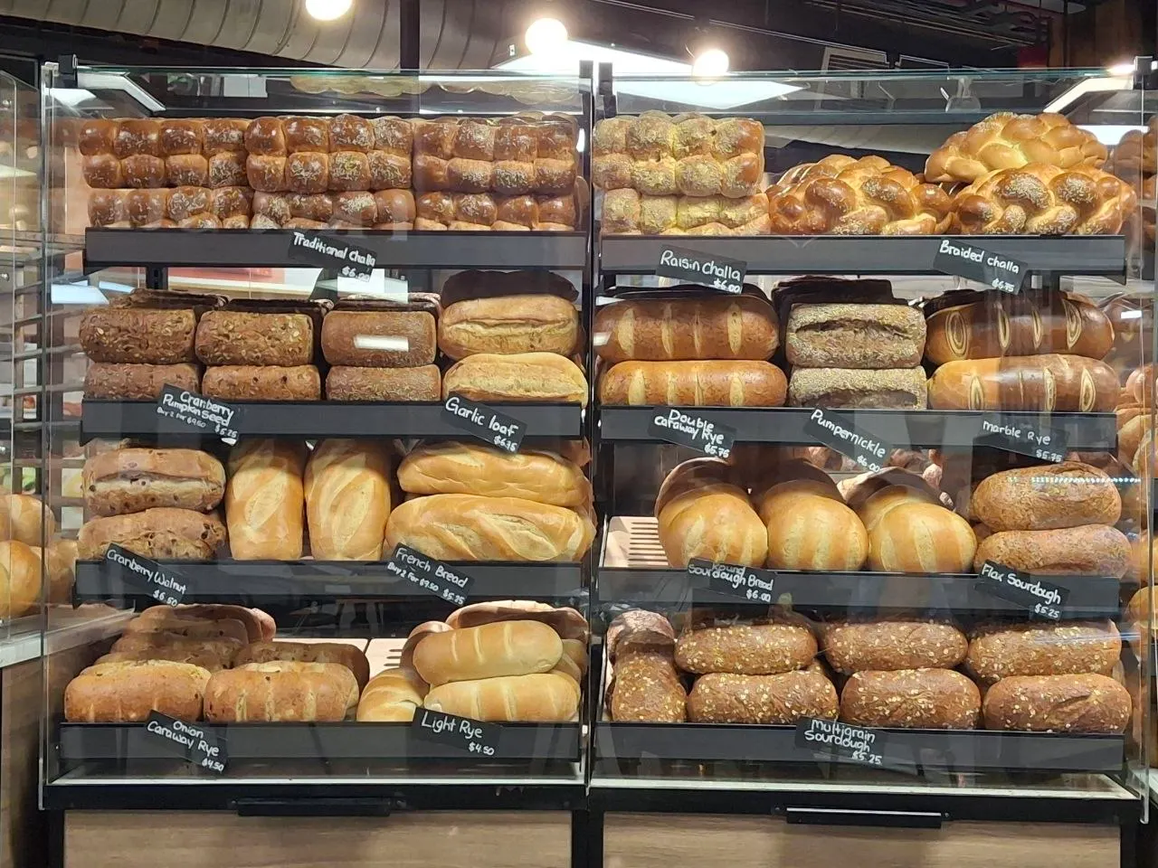 A bakery display case filled with various freshly baked loaves of bread, including challah, rye, sourdough, and French bread, each labeled with small black price tags.
