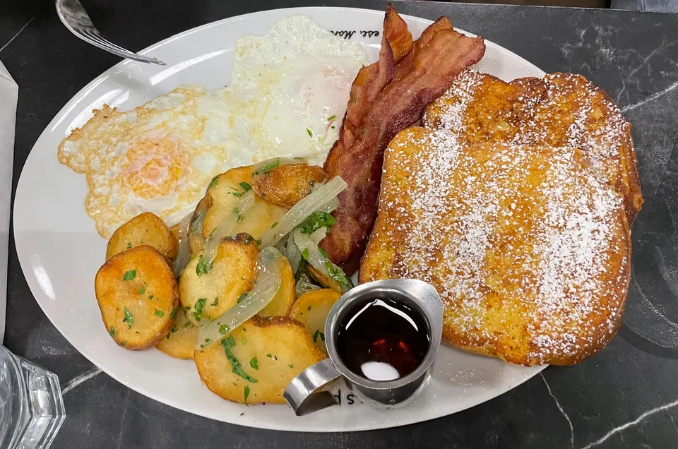 A breakfast plate featuring two fried eggs, home fries with onions and herbs, crispy bacon strips, and two thick slices of French toast dusted with powdered sugar, served with a small metal pitcher of syrup.