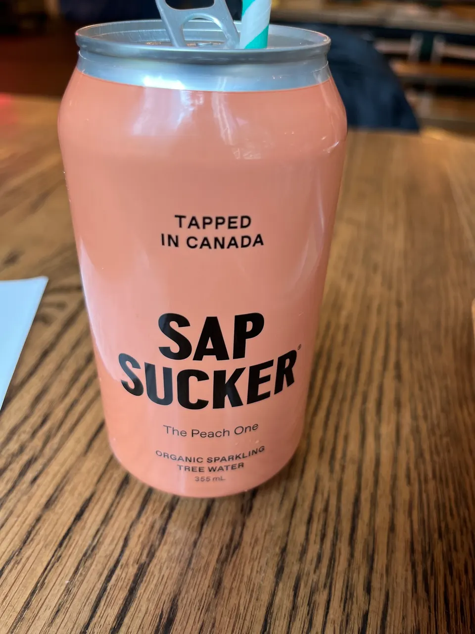 A peach-colored can of Sapsucker Organic Sparkling Tree Water, labeled 'The Peach One', sitting on a wooden table with a straw inserted.
