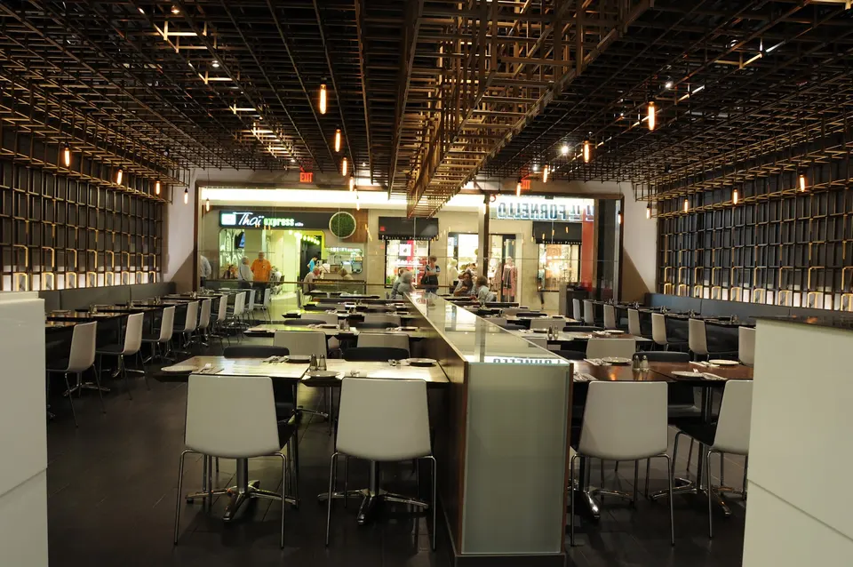 Interior view of IL FORNELLO restaurant at Bayview Village, featuring modern dining tables with white chairs under an intricate, dark wooden lattice ceiling with hanging warm lights.